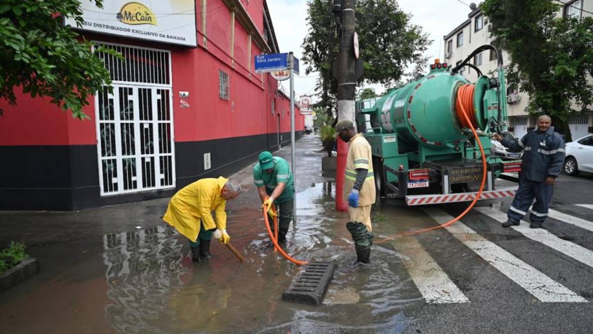 Equipe realizando drenagem e limpeza em Santos após fortes chuvas, com bombeamento e desobstrução de uma sarjeta na rua molhada em área urbana