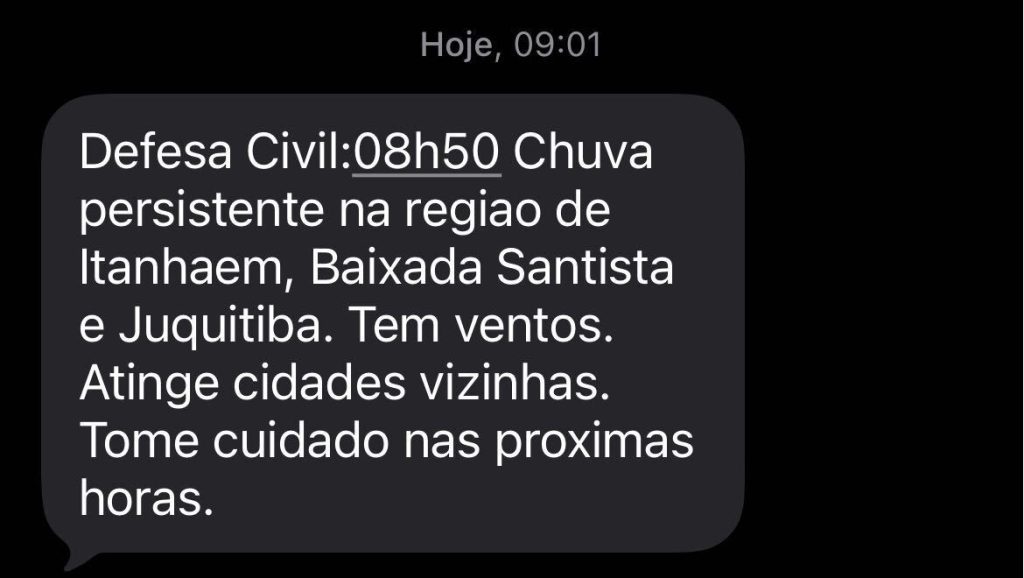 Aviso na tela com a mensagem “Defesa Civil: Chuva persistente na região de Itanhaém, Baixada Santista e Juquitiba”, com indicação de vento e orientação de cuidado nas próximas horas.