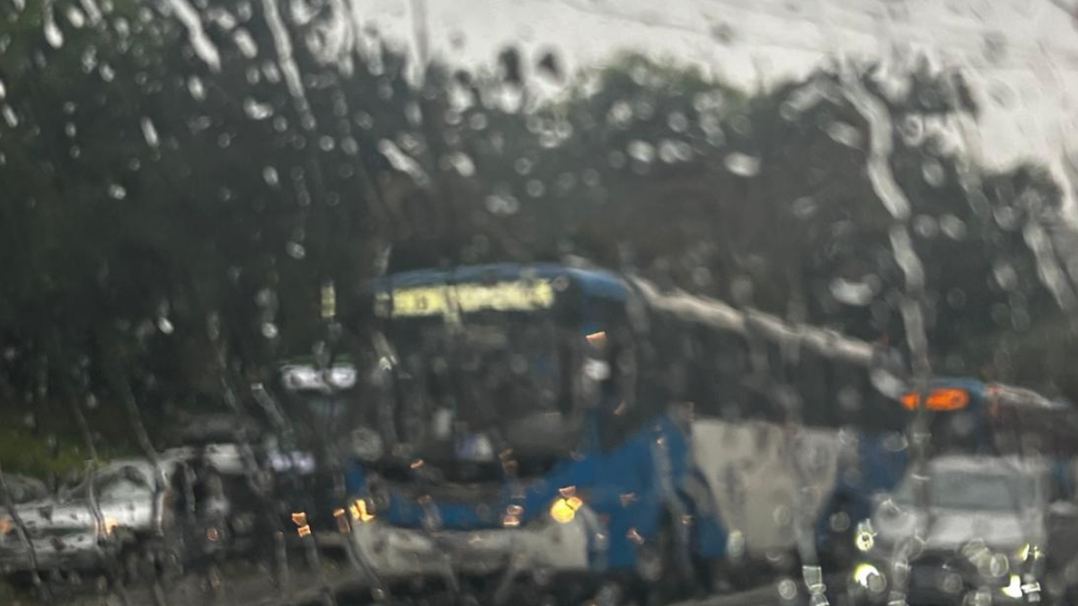 Vista em meio à chuva de uma estrada com carros e ônibus em movimento, com gotas no vidro e trânsito em destaque ao fundo.