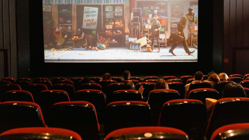 Semana do Cinema com destaque para uma sala de cinema lotada, poltronas vermelhas em primeiro plano e, ao fundo, a projeção de um filme em cartaz com cena urbana, em ambiente escuro.