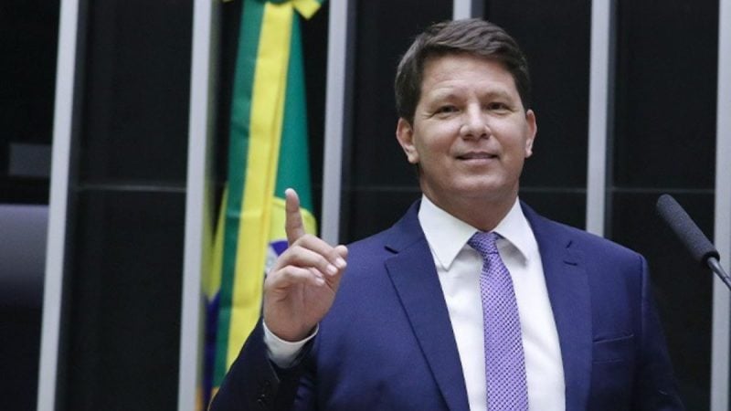 Mario Frias em foto de divulgação na Câmara dos Deputados, usando terno e gravata, ao lado da bandeira do Brasil e com microfone ao fundo.