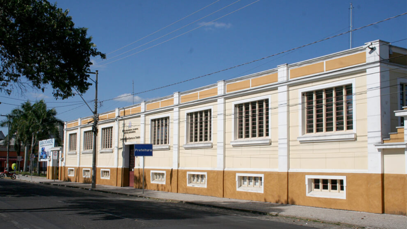 Fachada da Prefeitura de Amparo com dois andares com janelas emolduradas, faixa amarela e marrom e placas na entrada, visto de frente em uma rua sob céu azul.