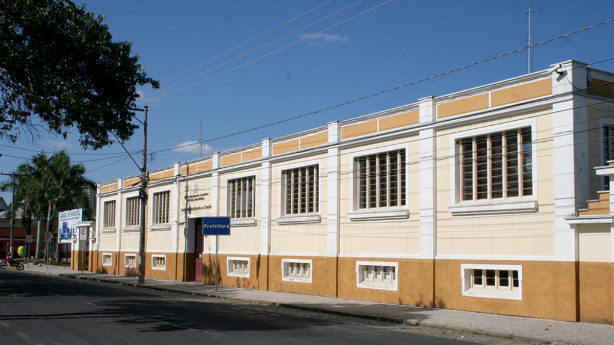 Fachada da Prefeitura de Amparo com dois andares com janelas emolduradas, faixa amarela e marrom e placas na entrada, visto de frente em uma rua sob céu azul.