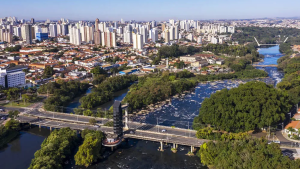 Vista aérea de Piracicaba com o rio atravessando o centro, grande ponte e áreas verdes, com bairros residenciais e prédios ao fundo ao entardecer.