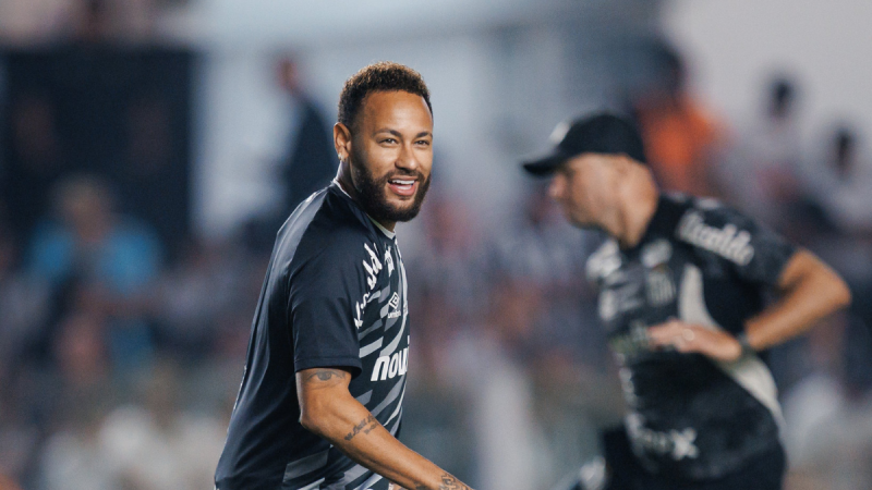 santos x atlético-mg: Neymar em campo durante partida entre Santos e Atlético-MG, com sorriso e expressão confiante, ao fundo torcedores e equipe técnica desfocados.