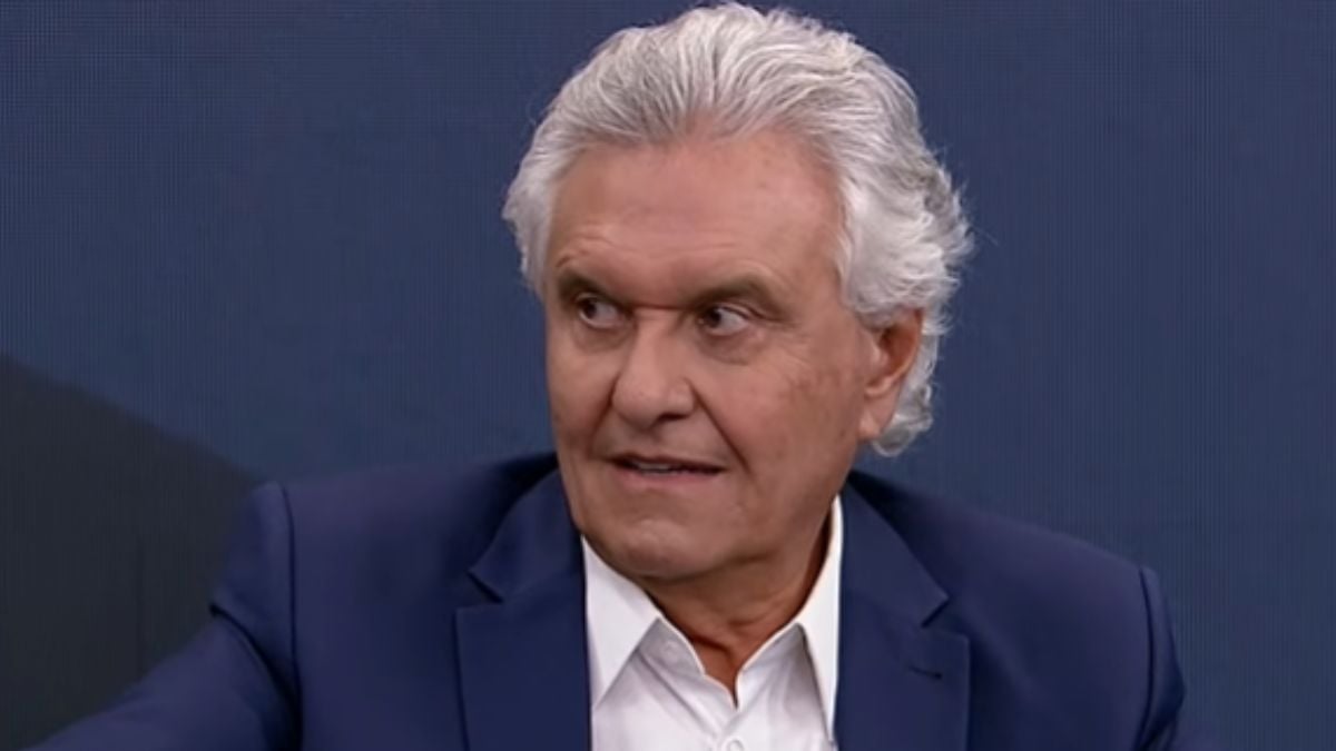 Ronaldo Caiado em entrevista, com expressão séria, em cenário político ao fundo, enquanto notícia informa que ele está internado em SP e cancelou a agenda de campanha