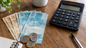 Lotofácil da Independência: cálculo do prêmio de R$ 300 milhões por mês, com calculadora, cédulas e moedas sobre mesa de madeira. Foto gerada por IA.