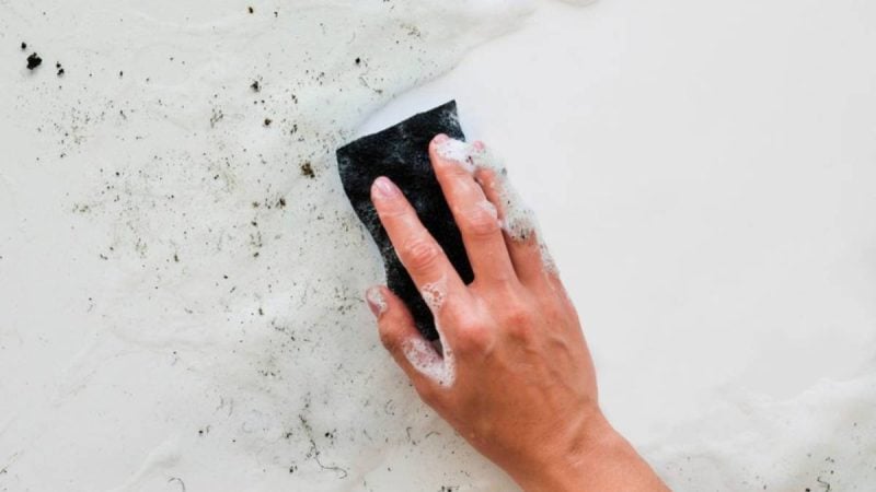 Mofo após chuva em uma parede: mãos limpando manchas escuras com esponja, mostrando acúmulo de fungos e necessidade de limpeza segura.