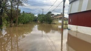 Inundação em Monte Mor: água alagando o entorno de uma propriedade com galpão de metal ao lado de árvores, postes e via próxima, com céu nublado.
