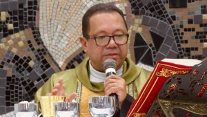 Padre falando ao microfone durante celebração religiosa, com livro litúrgico e paramentos em ambiente com mosaico; Diocese de Santos proíbe padre preso por suspeita de estupro de celebrar missas. Padre estupro Praia Grande