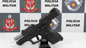 A Polícia Militar apresenta uma arma apreendida em mesa de investigação, com logotipos da Polícia Militar de São Paulo ao fundo, após investigação envolvendo dupla tentativa de homicídio em Atibaia.