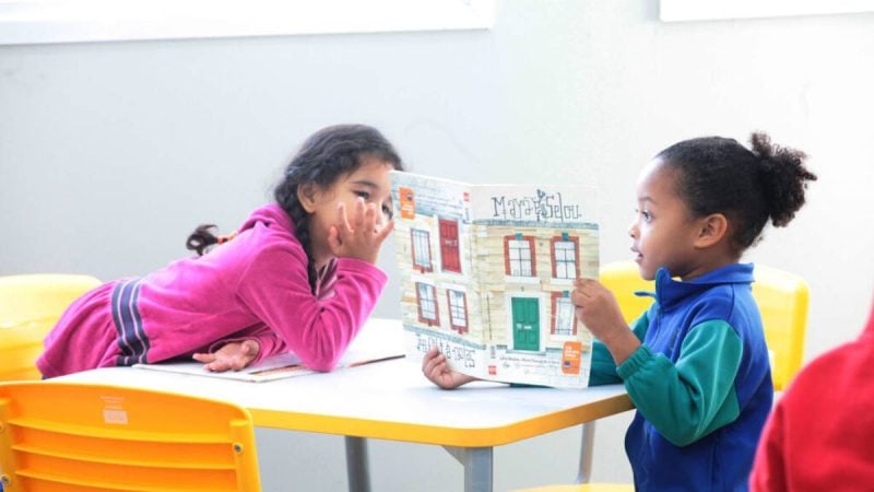 Crianças brincam e observam um livro em uma sala de aula, com mesa e material didático, ilustrando inscrições de crianças em Piracicaba para creche e pré-escola.