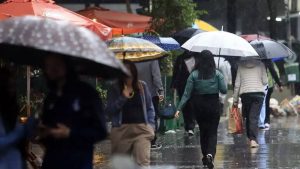 Pessoas caminhando em rua molhada durante chuva, em meio a guarda-chuvas e água acumulada, com alerta para alagamentos e deslizamentos na região bragantina