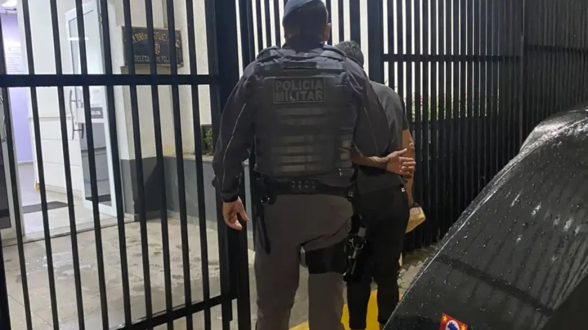 Policial militar conduz um homem algemado em frente a grades de uma delegacia, durante operação relacionada à tentativa de homicídio em Nazaré Paulista.