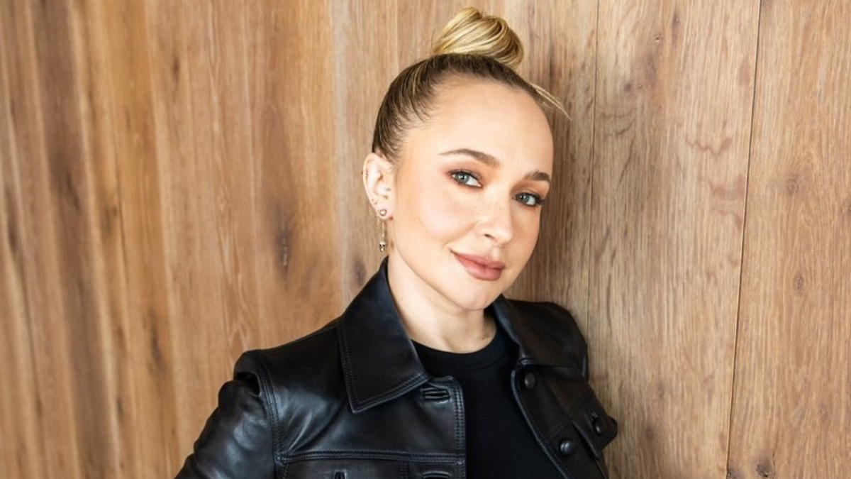 A atriz Hayden Panettiere de cabelo loiro preso em um coque e jaqueta de couro preta, sorrindo suavemente em frente a uma parede de madeira.