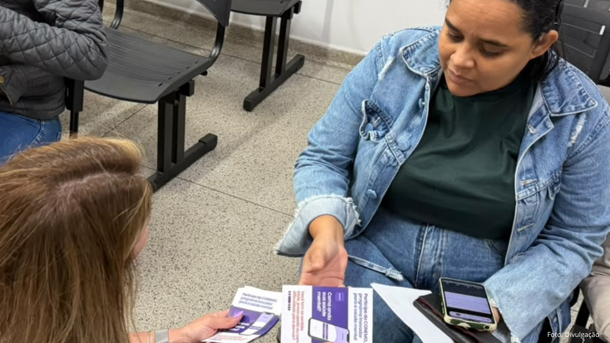 Mulher de jaqueta jeans segurando folhetos sobre saúde mental em uma UBS de Indaiatuba, enquanto conversa com outra mulher em uma sala com cadeiras pretas.