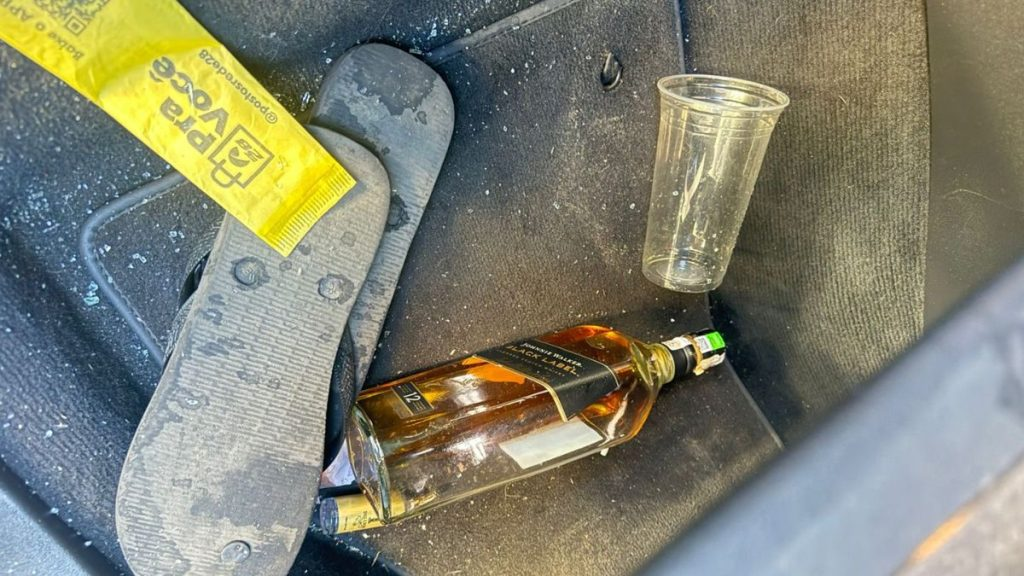 pais de motorista que matou coletor: Materiais apreendidos em investigação policial após atropelamento e morte de ciclista, com garrafa de bebida alcoólica, copo descartável e fita de isolamento amarelo com escrita da polícia sobre o piso.