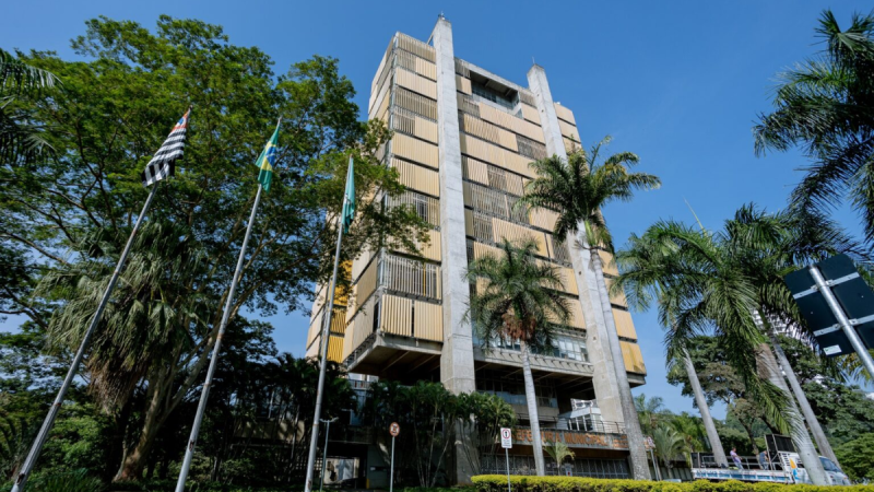 Fachada do prédio da Prefeitura Municipal de Piracicaba, com arquitetura moderna em concreto e brises amarelos nas janelas, cercado por palmeiras e árvores sob céu azul, com mastros exibindo as bandeiras do Brasil e do estado de São Paulo no canto esquerdo.