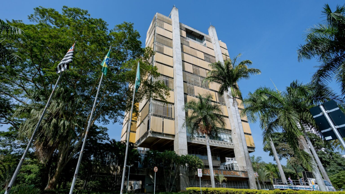 Fachada do prédio da Prefeitura Municipal de Piracicaba, com arquitetura moderna em concreto e brises amarelos nas janelas, cercado por palmeiras e árvores sob céu azul, com mastros exibindo as bandeiras do Brasil e do estado de São Paulo no canto esquerdo.