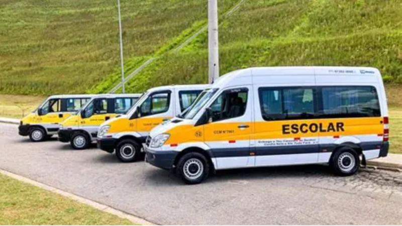 Ônibus escolares amarelos e brancos estacionados em fila na estrada, com a palavra “ESCOLAR” em destaque na lateral do veículo principal.