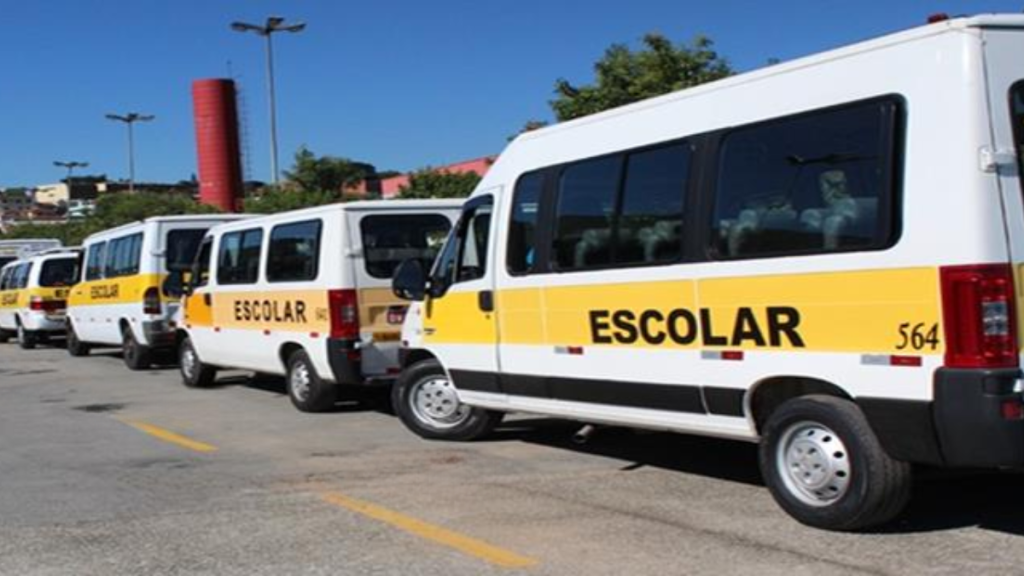Frota de vans escolares estacionadas em pátio, com faixas amarelas e a inscrição “ESCOLAR” na lateral, sob céu azul e iluminação diurna.