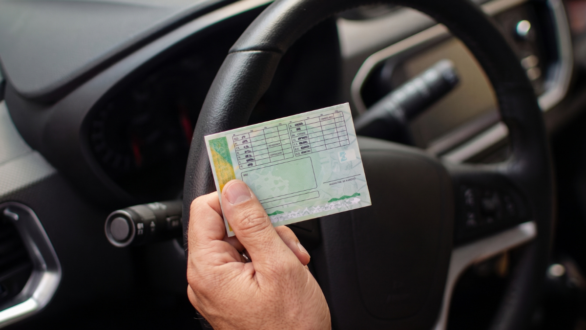 Foto em close da mão de um motorista segurando CNH junto ao volante, com o painel do veículo ao fundo, destacando dados impressos.