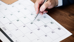 Pessoa usando uma caneta sobre um calendário, em ambiente de escritório, com foco