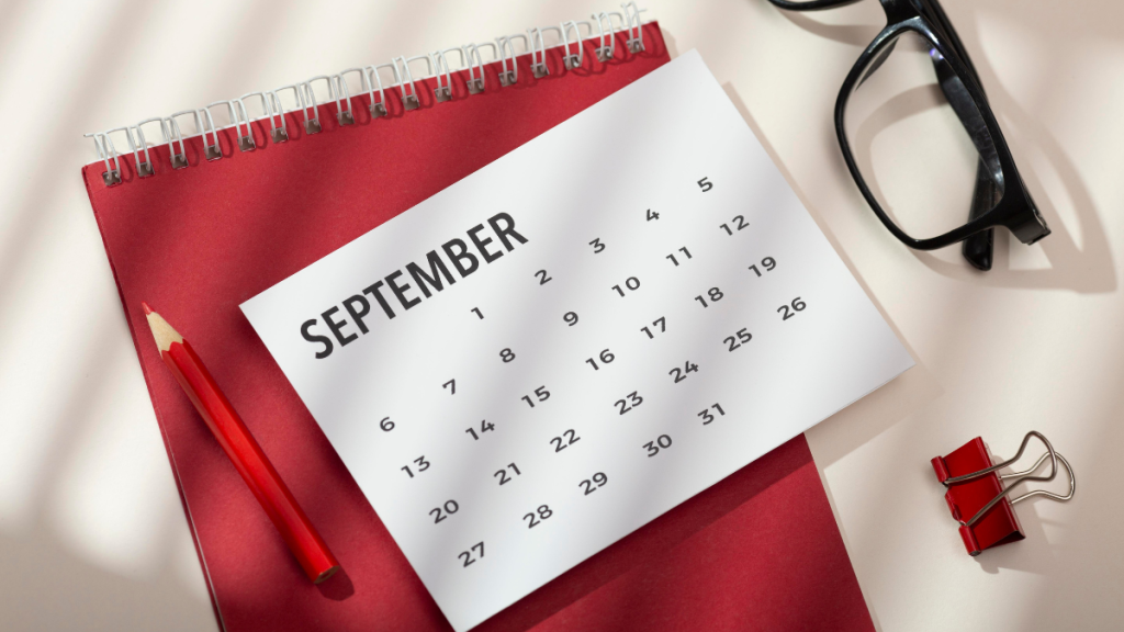 Calendário de setembro em destaque, com dias do mês numerados, em meio a papel vermelho, caneta e óculos sobre uma mesa clara, estilo planejador.