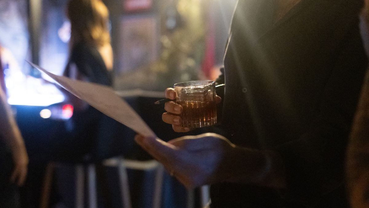 Negroni Week: mão ergue um copo de coquetel em um ambiente de bar com clima de speakeasy, ao fundo há garrafas e iluminação baixa.