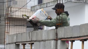 vagas de emprego em Praia Grande: Trabalhador em obra carregando materiais ao lado de um prédio em construção, com ambiente urbano que representa oportunidades locais. Praia Grande abre 128 vagas de emprego no PAT.