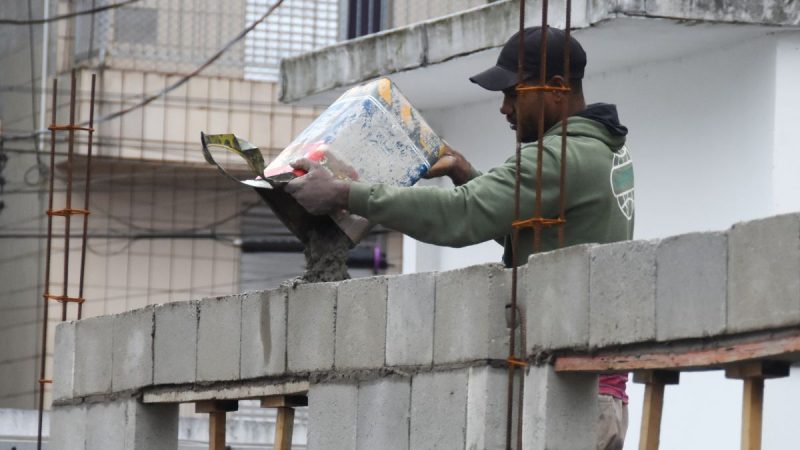 vagas de emprego em Praia Grande: Trabalhador em obra carregando materiais ao lado de um prédio em construção, com ambiente urbano que representa oportunidades locais. Praia Grande abre 128 vagas de emprego no PAT.