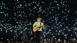 Ed Sheeran com violão no palco em um show ao vivo, cercado por um fundo escuro com muitas luzes brilhantes projetadas, enquanto o público observa.