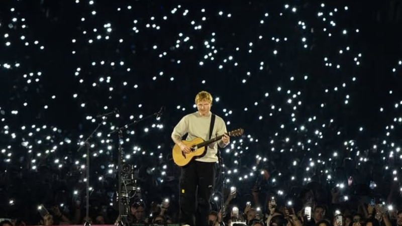 Ed Sheeran com violão no palco em um show ao vivo, cercado por um fundo escuro com muitas luzes brilhantes projetadas, enquanto o público observa.