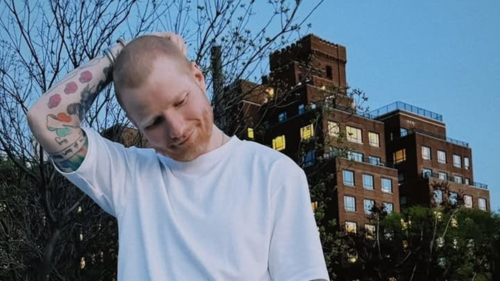 Ed Sheeran com tatuagens no braço usa camiseta branca e inclina a cabeça, em ambiente externo com árvores ao fundo e prédios de tijolos ao céu azul.