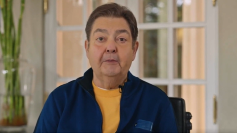 O apresentador de televisão Fausto Silva (Faustão), um homem adulto de cabelos castanhos curtos, olha diretamente para a câmera com uma expressão séria e neutra. Ele veste uma blusa com zíper azul-marinho sobre uma camiseta amarela. Ao fundo, há uma janela ou porta de vidro dividida em quadrículos e, à esquerda, um vaso transparente com hastes de bambu-da-sorte.