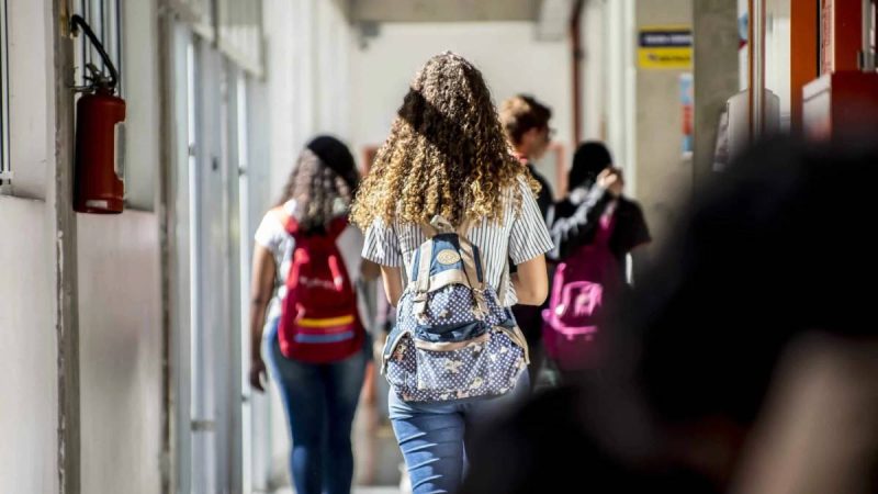 Pessoas caminhando em corredor de instituição de ensino, com mochilas e alunos ao fundo, reforçando a abertura de vagas do Centro Paula Souza para ensino médio, técnico e graduação.