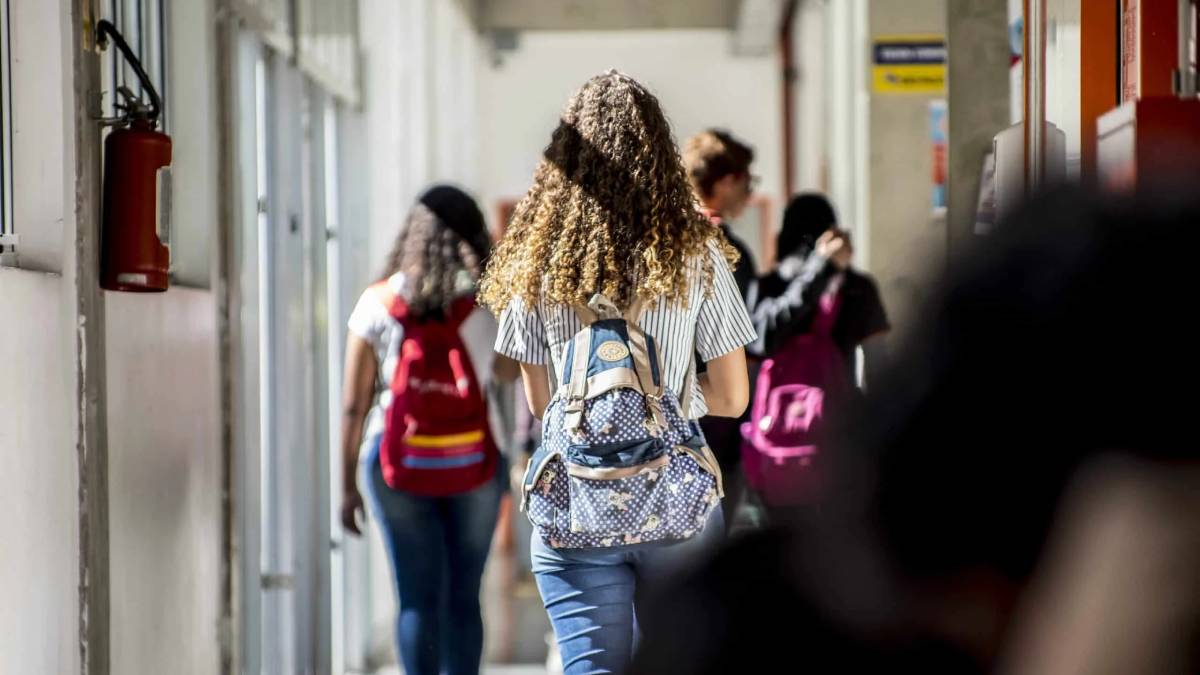 Pessoas caminhando em corredor de instituição de ensino, com mochilas e alunos ao fundo, reforçando a abertura de vagas do Centro Paula Souza para ensino médio, técnico e graduação.