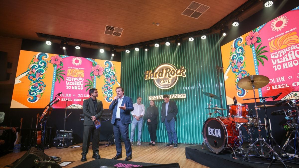 Palco do Estação Verão Show com telões coloridos, bateria e músicos durante apresentação na Praia Grande, divulgando a programação do evento 2027.
