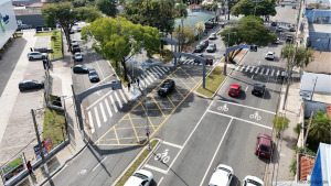 Vista aérea de um cruzamento em Indaiatuba com faixas de pedestres, sinalização amarela de cruzamento, ciclovia demarcada e semáforos instalados em pórticos metálicos.