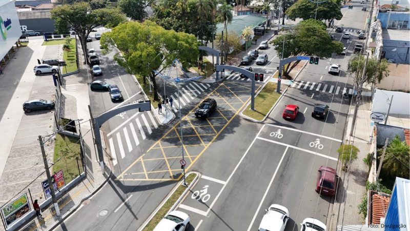 Vista aérea de um cruzamento em Indaiatuba com faixas de pedestres, sinalização amarela de cruzamento, ciclovia demarcada e semáforos instalados em pórticos metálicos.
