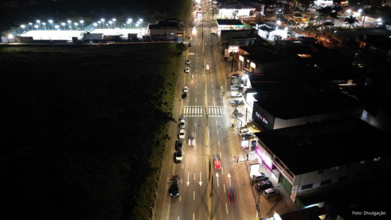 Foto aérea noturna mostrando a iluminação pública de uma avenida em Indaiatuba. A pista exibe marcações viárias, carros em movimento e edifícios comerciais iluminados ao longo da via.