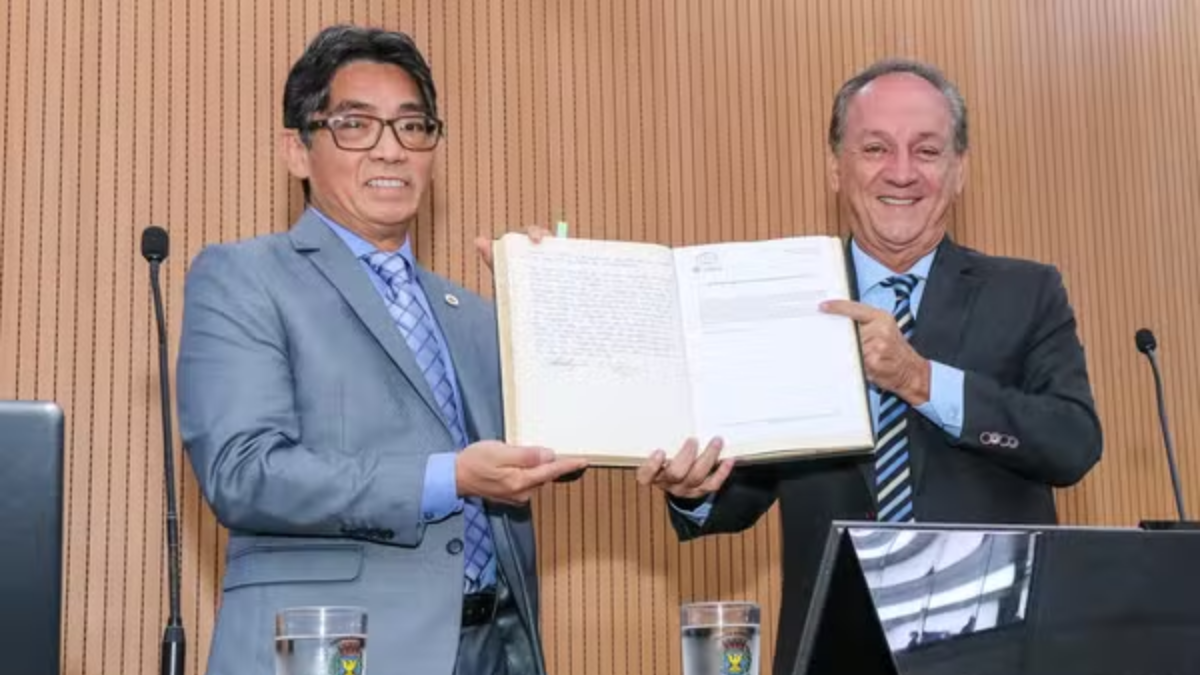 Na esquerda, o novo vereador Tak com presidente da Câmara, Luiz Rossini em um auditório, sorrindo e segurando sua posse durante uma cerimônia de assinatura