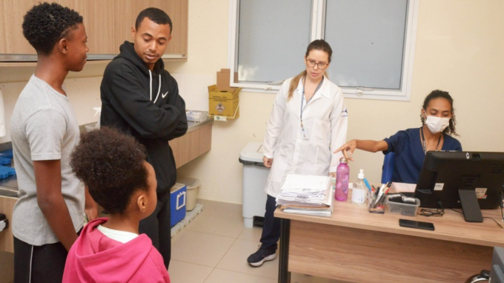 Ambiente de atendimento em uma clínica a cidade de Campinas: com mesa de trabalho, computador e materiais, enquanto outras pessoas aguardam usando máscara.