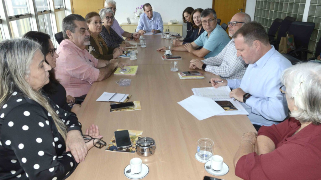 Reunião de Helinho Zanatta com membros da Academia Piracicabana de Letras, secretários e vereador ao redor de uma mesa de escritório, analisando documentos sobre a nova sede no bairro Jaraguá.
