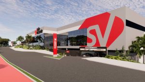 Vista externa de um prédio comercial moderno com fachada branca e grande logotipo vermelho “SV”, área de estacionamento, caminho com faixa vermelha e entrada sinalizada, em ambiente urbano.