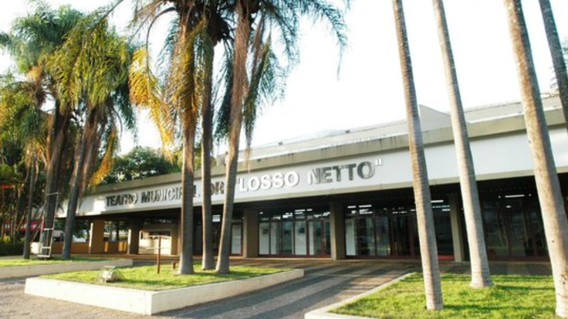Vista externa da fachada moderna e térrea do Teatro Municipal Dr. Losso Netto em Piracicaba, destacando o letreiro na parte superior da entrada e várias palmeiras altas espalhadas pelos canteiros de grama e caminhos pavimentados em frente ao prédio.