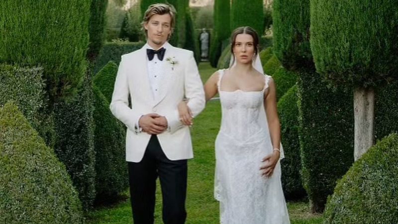 Millie: Millie Bobby Brown posa com Jake Bongiovi em um jardim com arbustos e caminho ajardinado, usando traje formal branco em foto de casal ao ar livre.