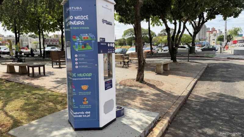 Estação de hidratação vertical instalada no Parque Ecológico de Indaiatuba. A estrutura metálica é branca e azul, decorada com infográficos e frases informativas sobre a água. Na parte frontal, há um dispensador de água e, na base ao chão, um bebedouro para pets. Ao fundo, veem-se árvores, bancos do parque, ruas e veículos.
