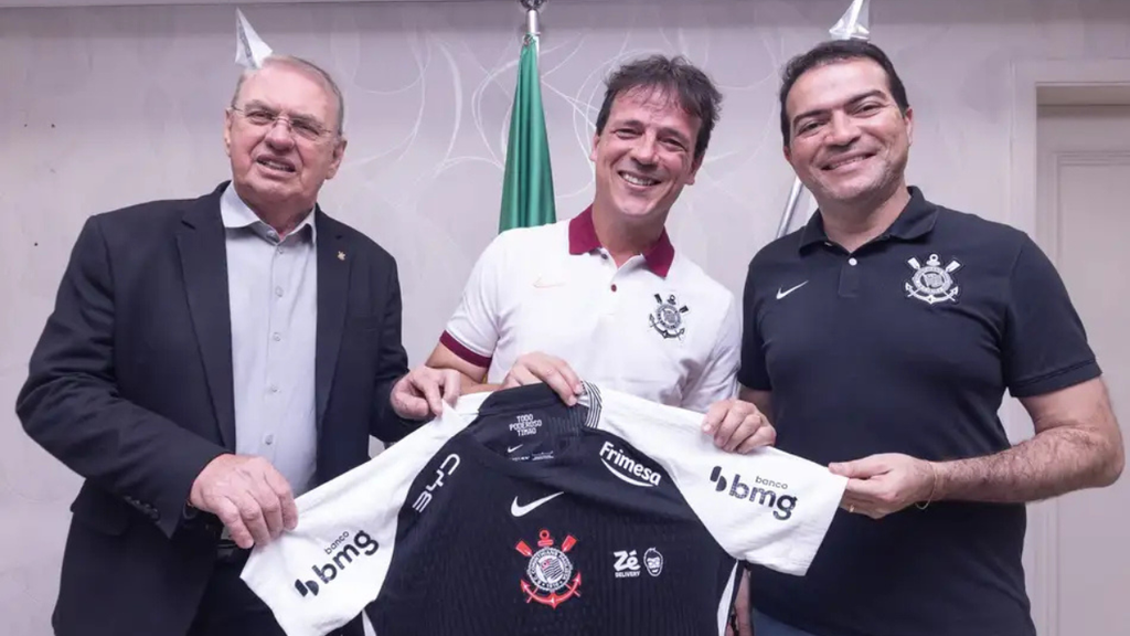 Fernando Diniz Corinthians com Osmar Stabile e Marcelo Paz em foto de apresentação, segurando camisas do Corinthians, ao lado de bandeira e em ambiente institucional.