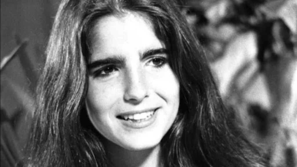 Fotografia em preto e branco com plano aproximado (rosto) da atriz brasileira Malu Mader na juventude, sorrindo de forma suave e olhando ligeiramente para a direita. Ela tem cabelos escuros, longos e soltos, com sobrancelhas marcantes e espessas. O fundo está desfocado, com folhagens e iluminação natural.